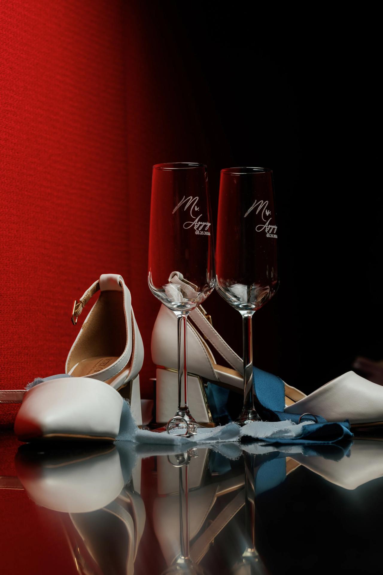 Wedding shoes and two champagne flutes styled together on a red background, with a blue fabric on the table. Wedding photography at Bellworks, Holmdel, NJ.
