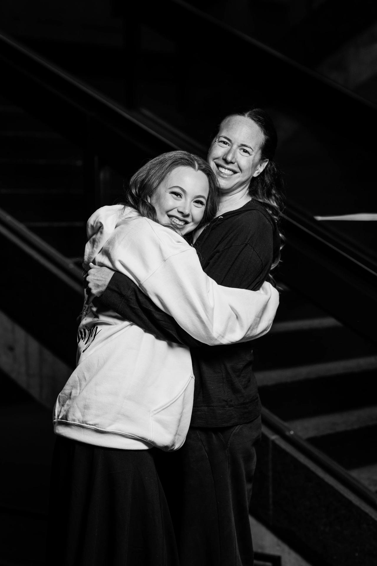 Black-and-white dance-floor moment: two women smiling in a close hug, framed by dark architectural lines and strong shadow lighting. Wedding photography at Bellworks, Holmdel, NJ.