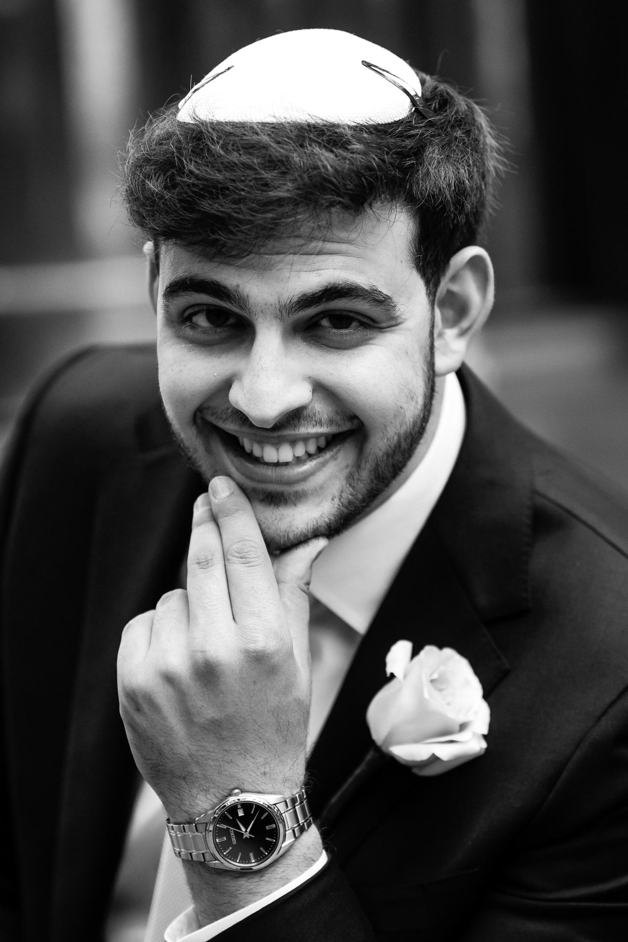 Black-and-white groom portrait in a kippah smiling and holding up a peace sign, with a boutonnière and watch visible. Wedding photography at Bellworks, Holmdel, NJ.