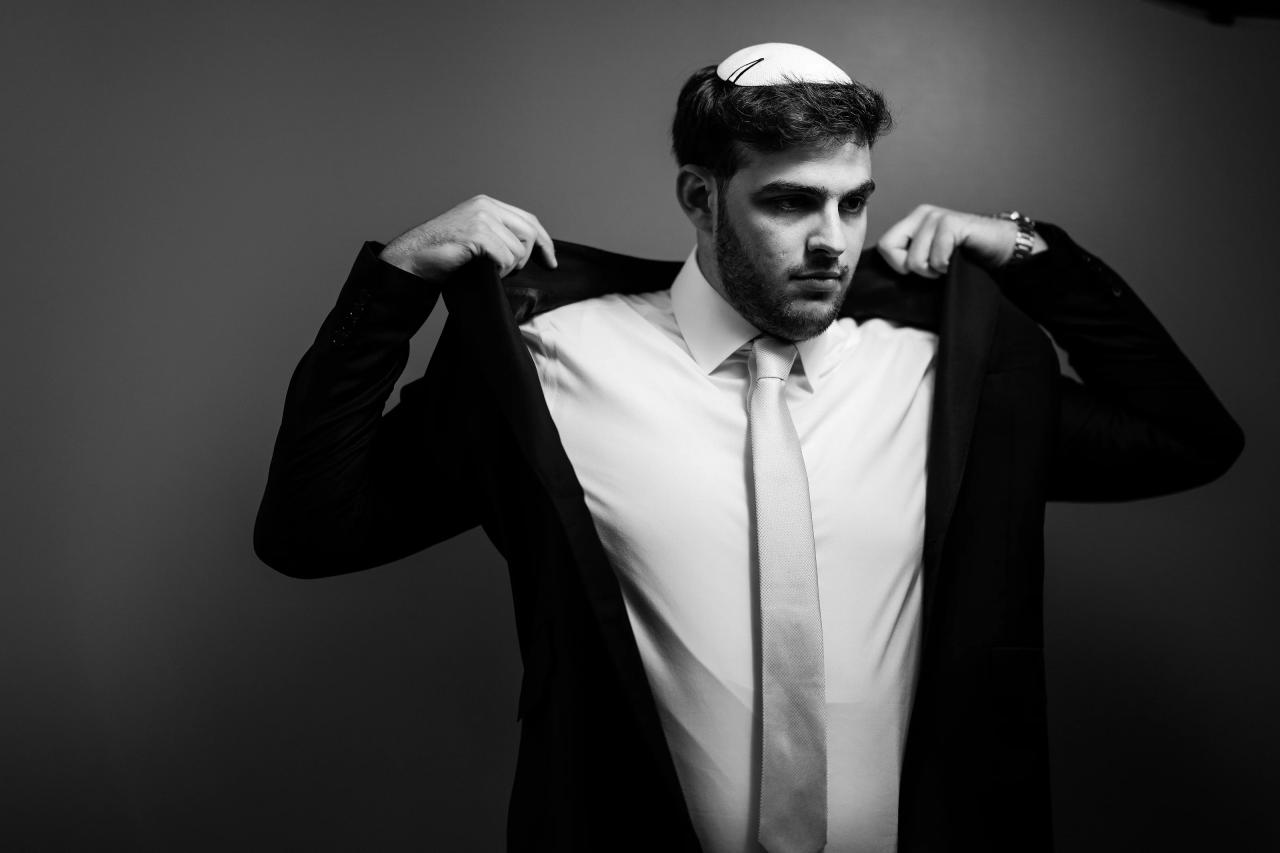 Black-and-white groom portrait indoors, looking at the camera while adjusting his suit lapels, with a plain dark backdrop. Wedding photography at Bellworks, Holmdel, NJ.