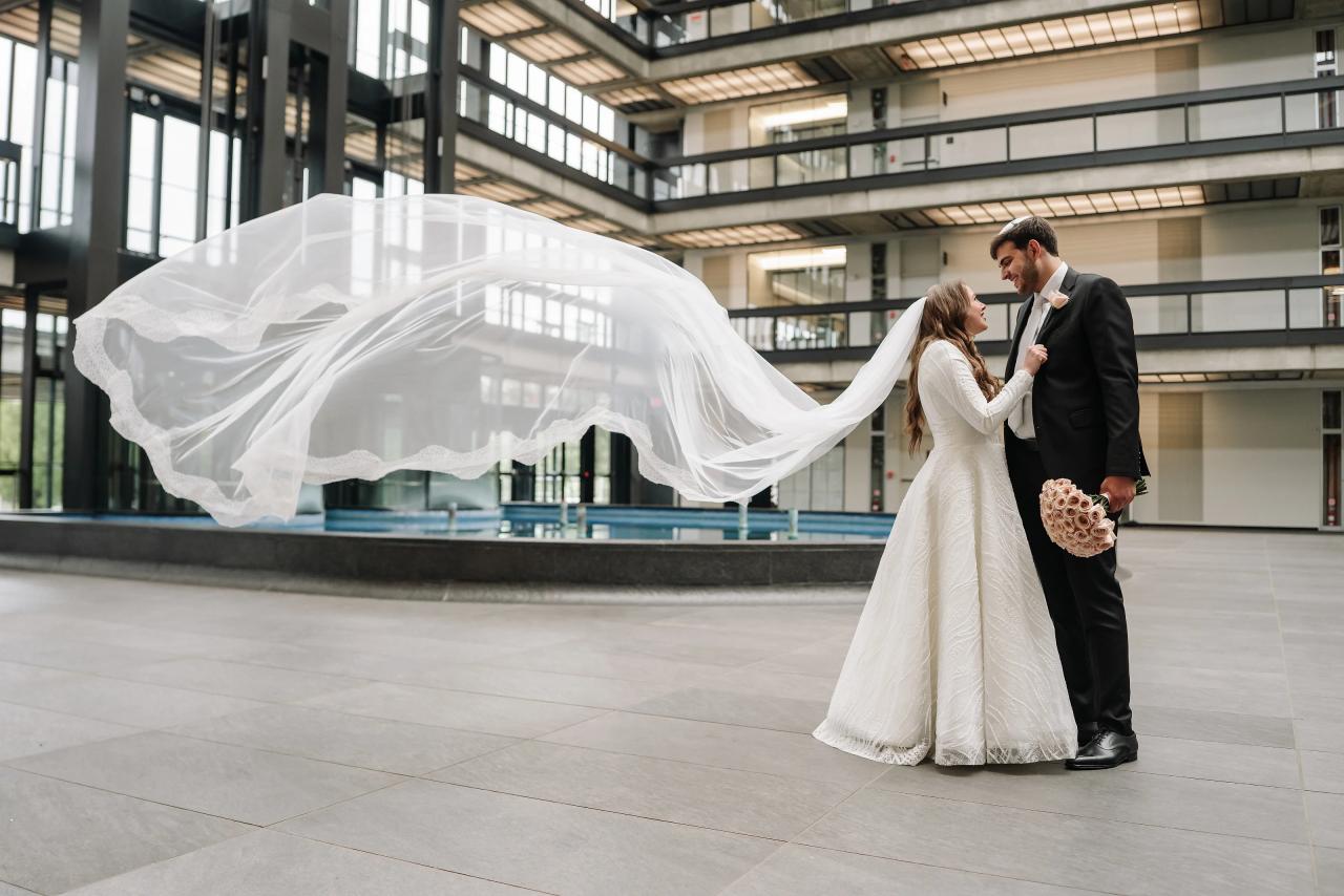 Newlyweds in a modern hall holding hands as the bride’s veil billows dramatically in the air. Wedding photography at Bellworks, Holmdel, NJ.