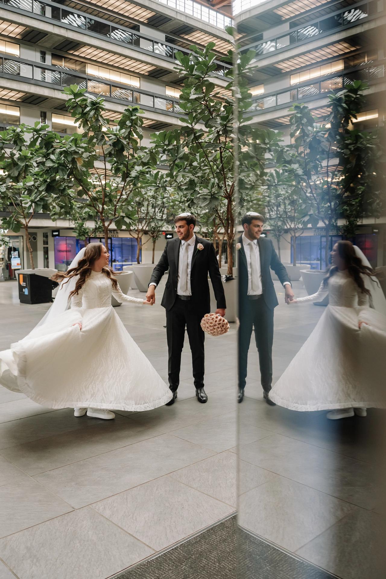 Bride and groom walking hand-in-hand with flowing wedding dresses, captured with playful motion in a modern indoor atrium with greenery. Wedding photography at Bellworks, Holmdel, NJ.