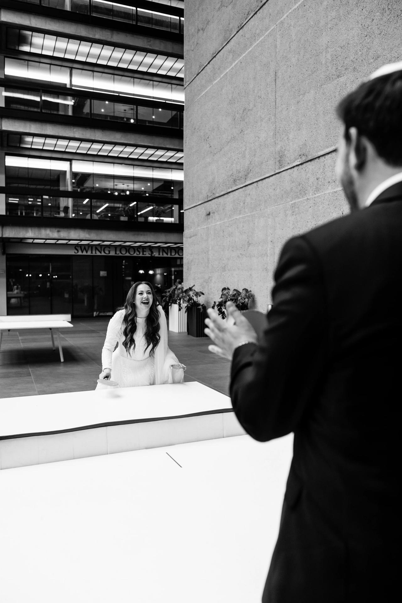 Black-and-white candid wedding photo of a bride laughing on a raised platform while guests react nearby, with hands reaching toward the moment. Wedding photography at Bellworks, Holmdel, NJ.