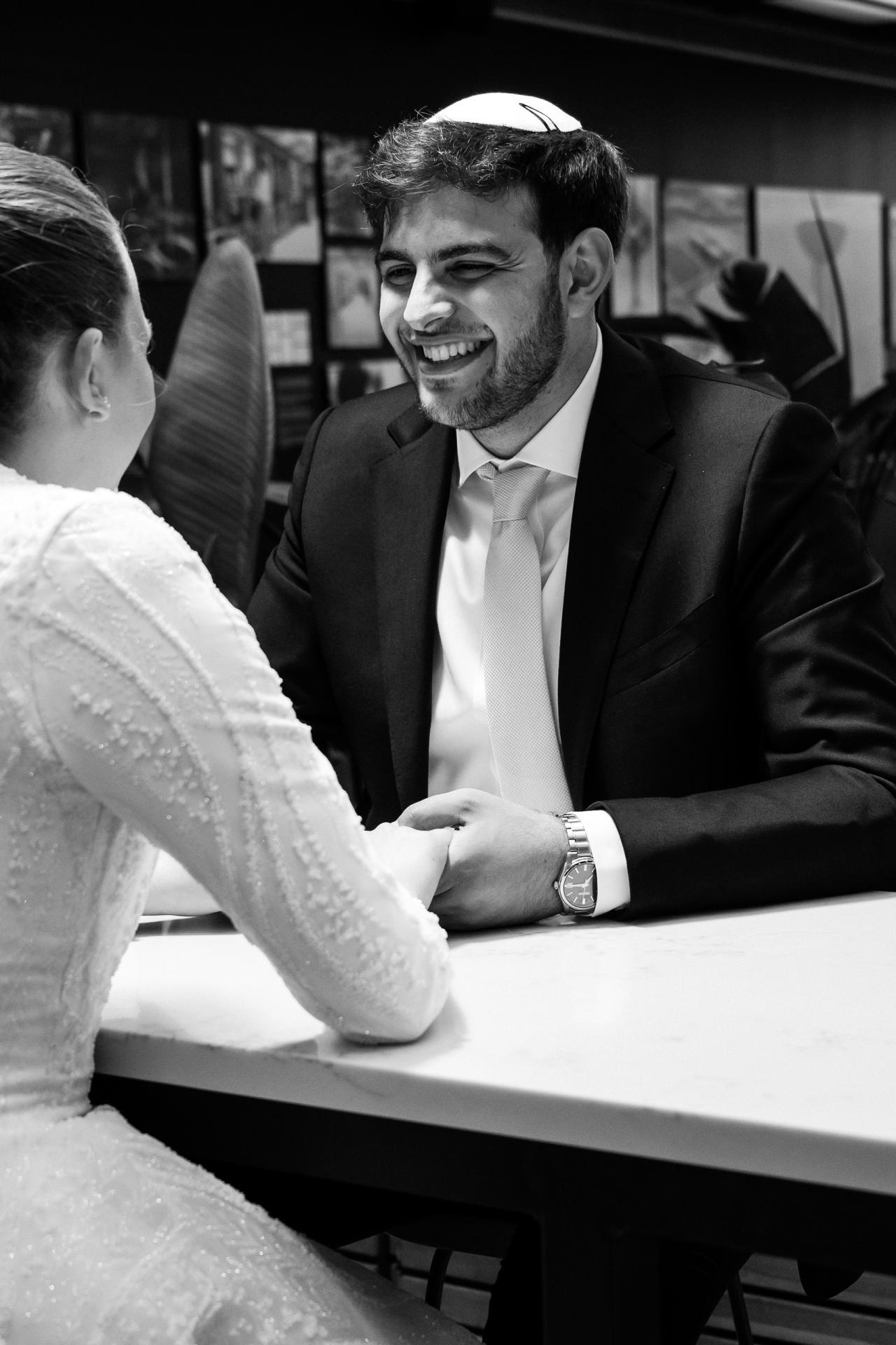 Black-and-white wedding reception candid: groom in a suit laughing while seated across from a guest at a table. Wedding photography at Bellworks, Holmdel, NJ.