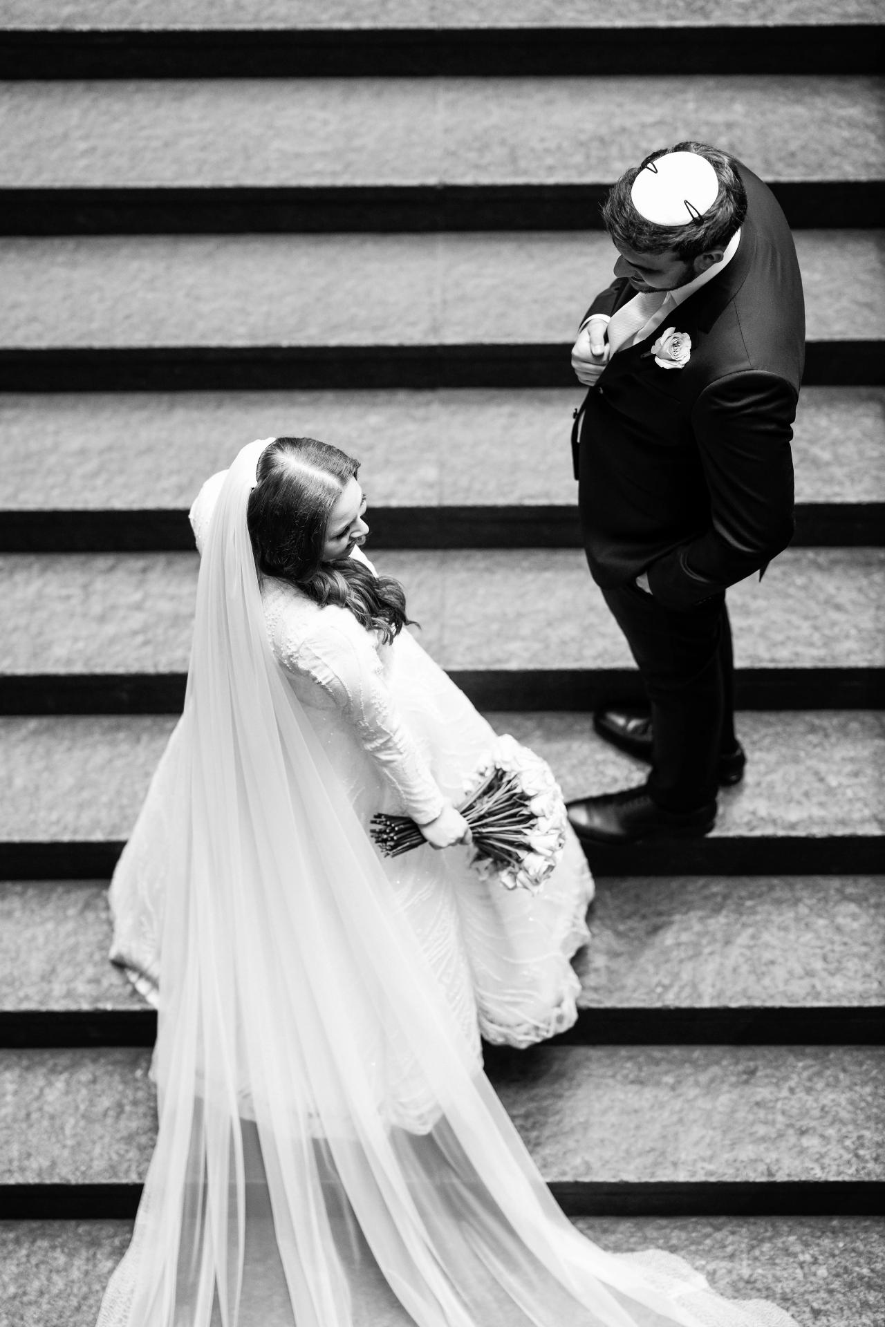 Black-and-white ceremony moment: a bride in a veil looks downward while holding her bouquet, standing close beside a groom in a black suit near a staircase. Wedding photography at Bellworks, Holmdel, NJ.