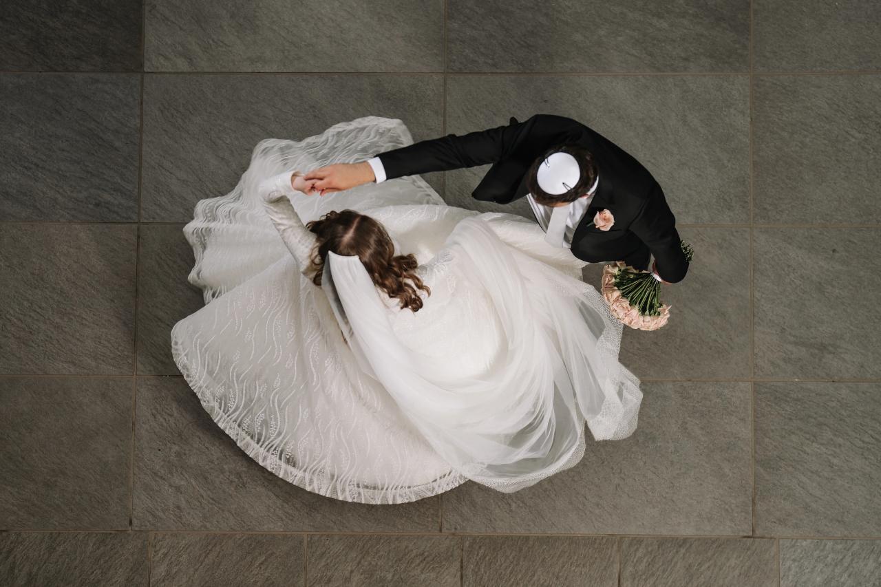 Top-down view of a bride and groom embracing indoors, bride’s flowing white dress and veil spread across a gray stone floor, groom holding a small bouquet. Wedding photography at Bellworks, Holmdel, NJ.