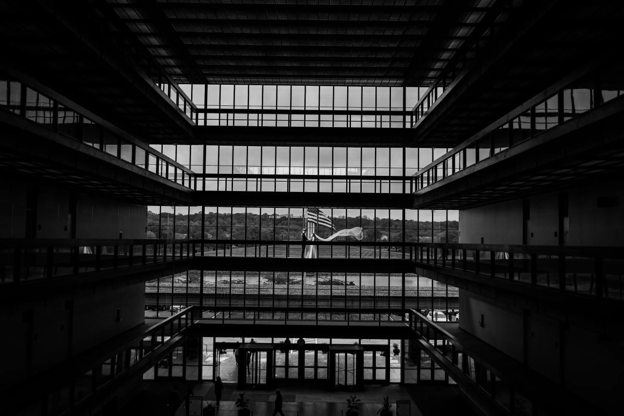 Black-and-white wide interior photograph with strong architectural lines and a small couple centered in the atrium, creating a graphic, solemn mood. Wedding photography at Bellworks, Holmdel, NJ.