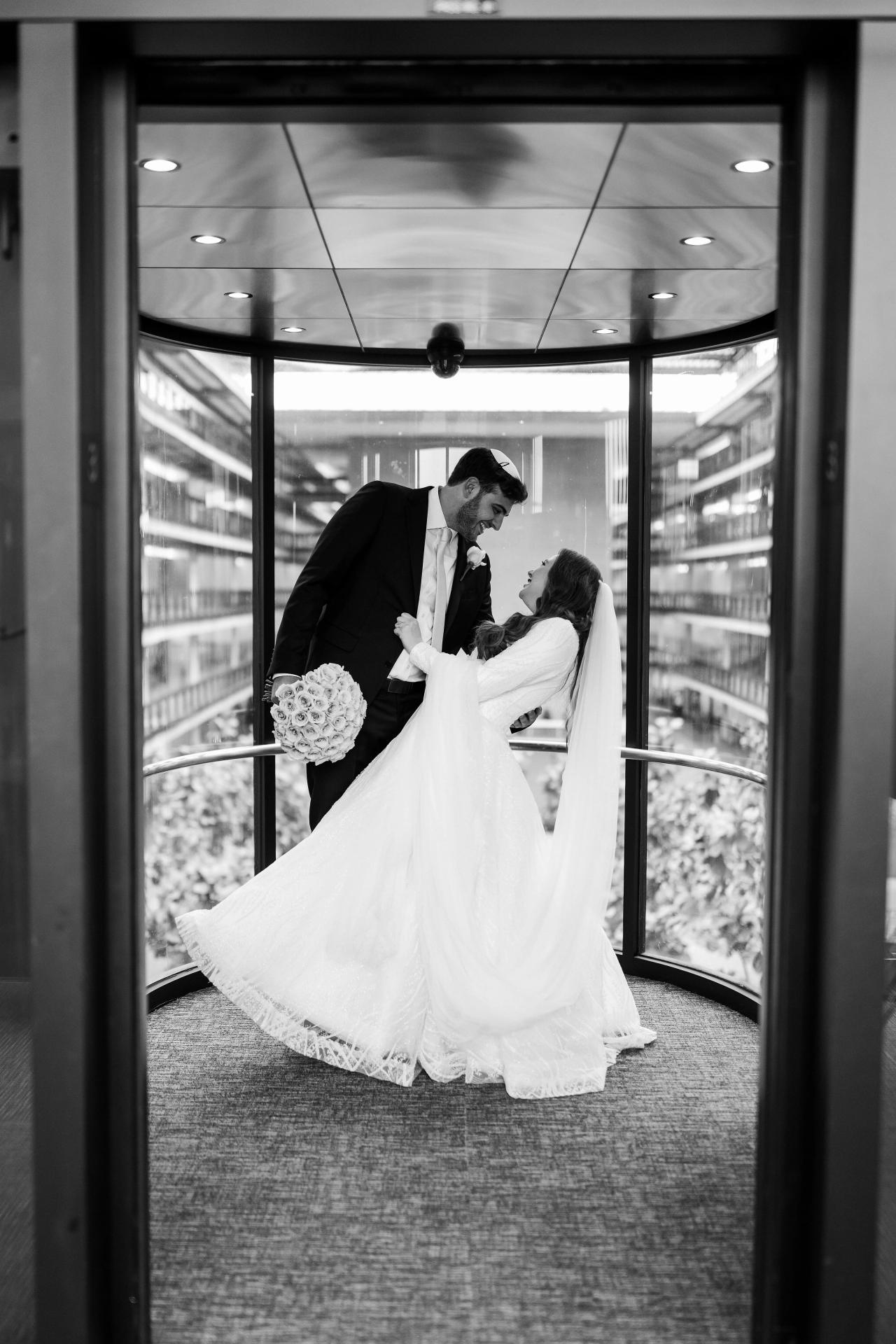 Black-and-white photo of a bride and groom sharing an intimate close embrace framed by glass doors, bride’s flowing veil extended as they lean in. Wedding photography at Bellworks, Holmdel, NJ.