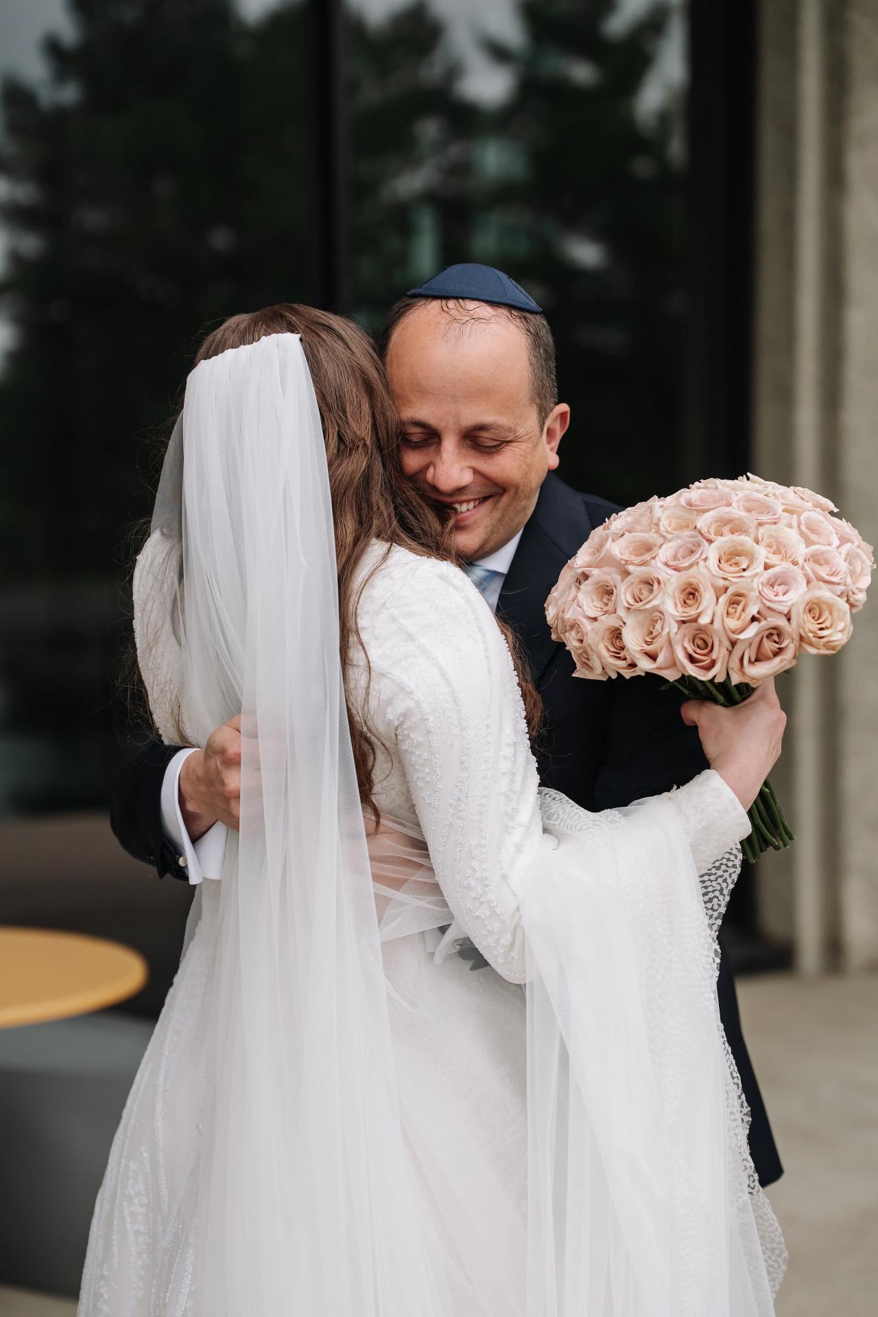 Bride and groom in a close embrace outdoors at a wedding ceremony, bride’s veil covering part of her face and the groom smiling while holding a bouquet of pale pink roses. Wedding photography at Bellworks, Holmdel, NJ.