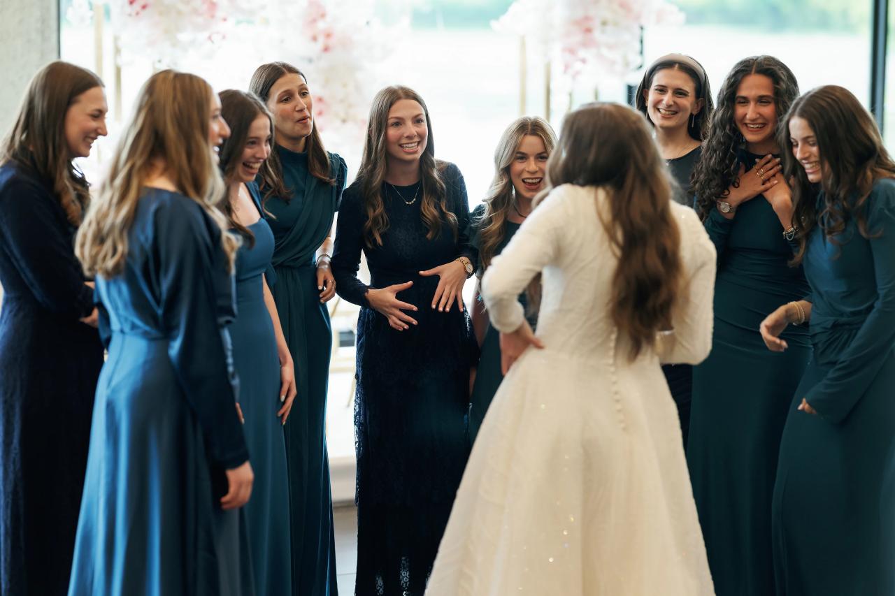 Wedding reception photo of a bride in a white gown surrounded by smiling women in dark blue dresses during a lively celebration in a bright hall with window light. Wedding photography at Bellworks, Holmdel, NJ.