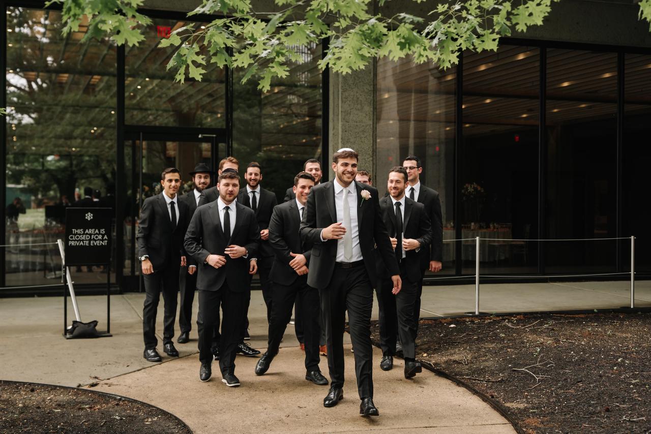 Group of suited men in formalwear walking outside along a curved walkway near a glass building entrance, with trees overhead. Wedding photography at Bellworks, Holmdel, NJ.