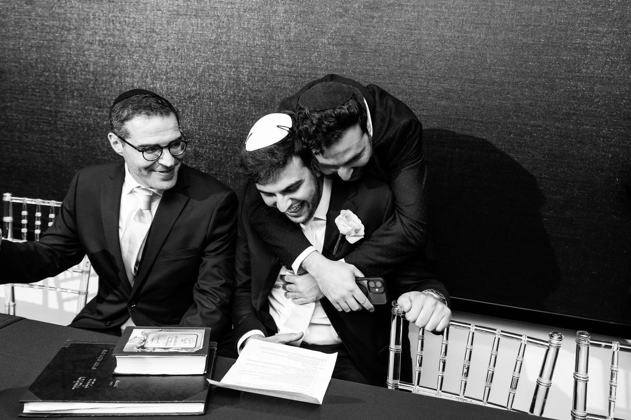 Black-and-white candid moment of a couple and close family leaning in and laughing at a reception table with papers and an ornate silver candelabra in the foreground. Wedding photography at Bellworks, Holmdel, NJ.