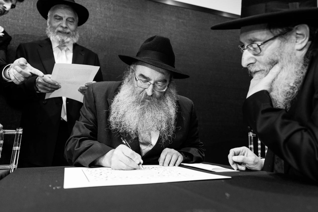 Black-and-white photo of a man seated in prayer attire, leaning forward to write or sign during a formal moment, with two other men seated behind him. Wedding photography at Bellworks, Holmdel, NJ.