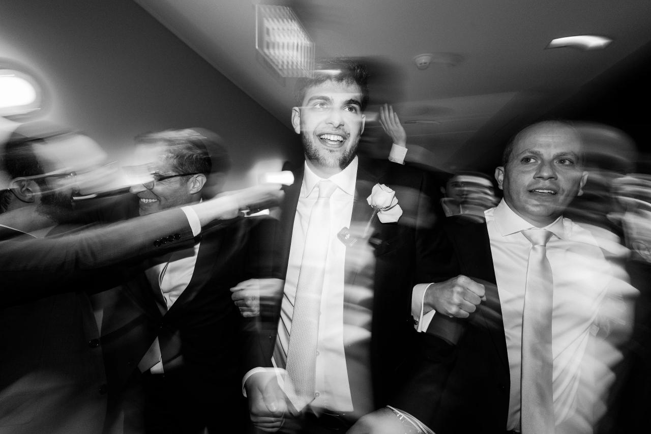 Black-and-white dance-floor photo of a smiling groom in a suit, with motion blur streaking around him and guests on either side raising hands and celebrating. Wedding photography at Bellworks, Holmdel, NJ.