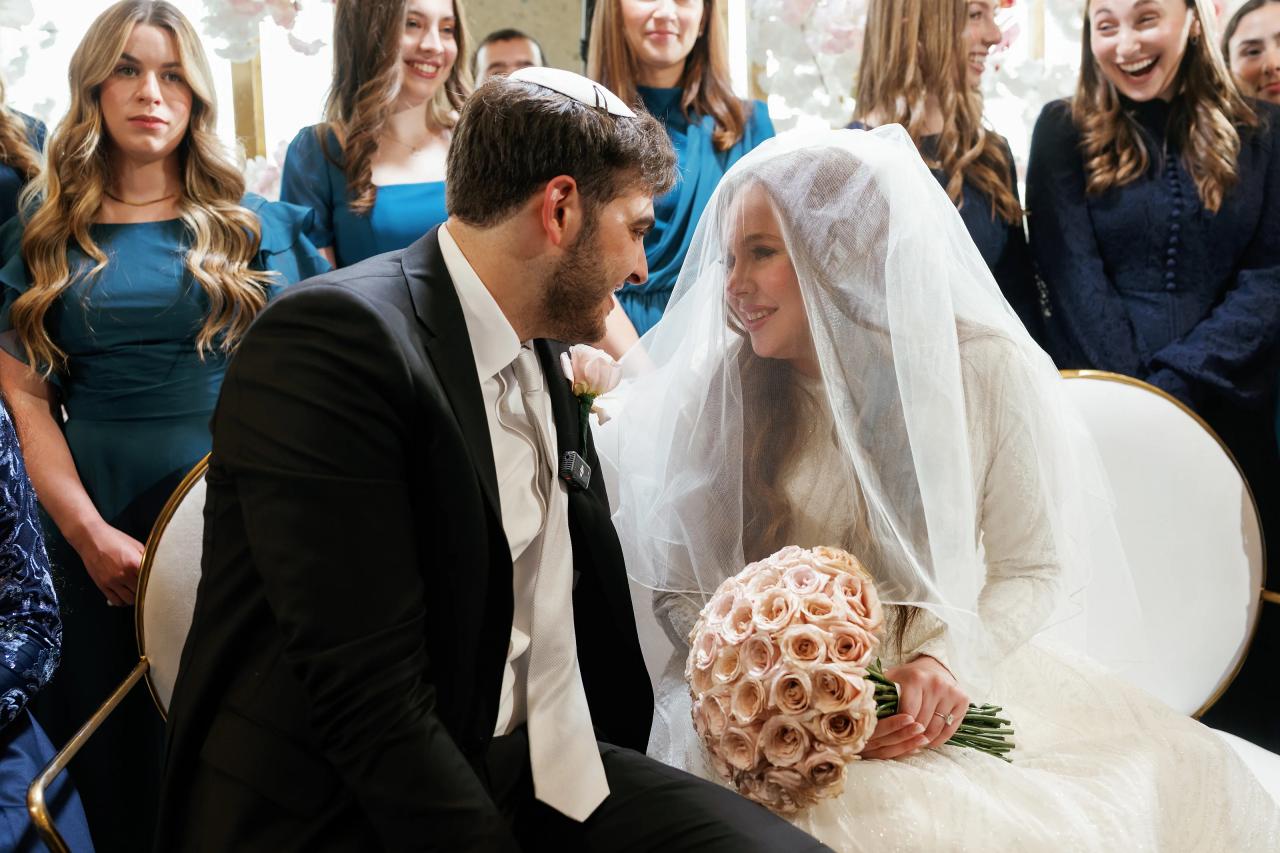 Groom gently leans in toward the bride during badeken as she smiles under a translucent veil, holding a bouquet; seated ceremony moment with guests softly blurred behind. Wedding photography at Bellworks, Holmdel, NJ.