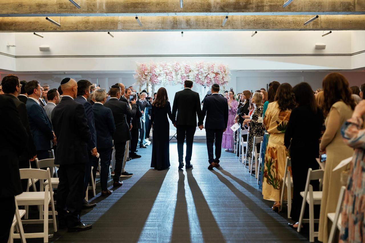 Guests standing along a wide aisle facing a floral chuppah at the front of the ceremony space, with soft indoor lighting and neutral walls. Wedding photography at Bellworks, Holmdel, NJ.