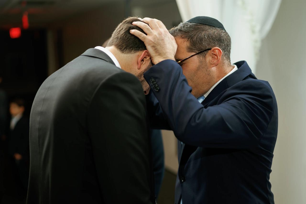 Two men in suits embrace closely, foreheads and hands touching in a heartfelt moment at a formal event. Wedding photography at Bellworks, Holmdel, NJ.