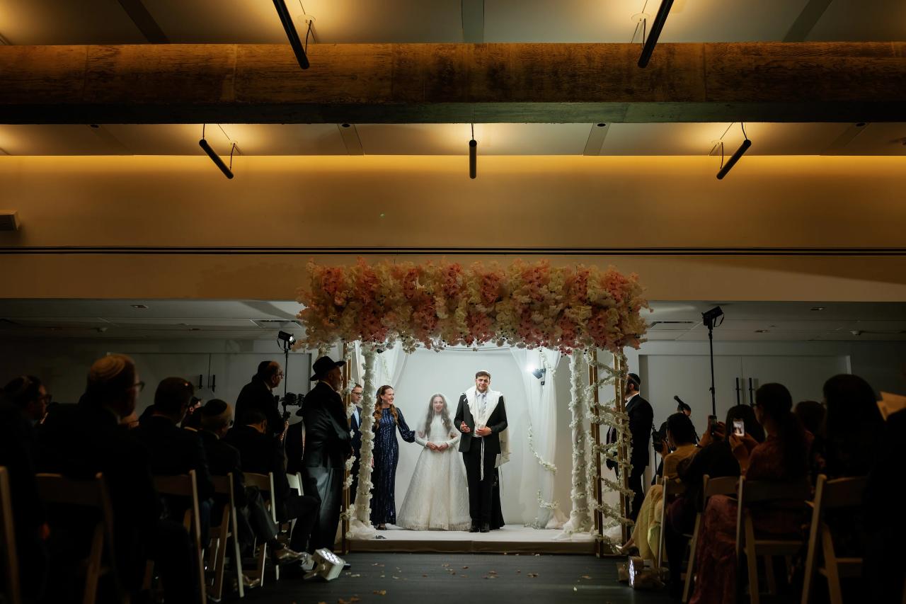 Wide ceremony photo of a wedding couple standing under a floral chuppah at the center aisle, guests seated on both sides in warm indoor lighting. Wedding photography at Bellworks, Holmdel, NJ.