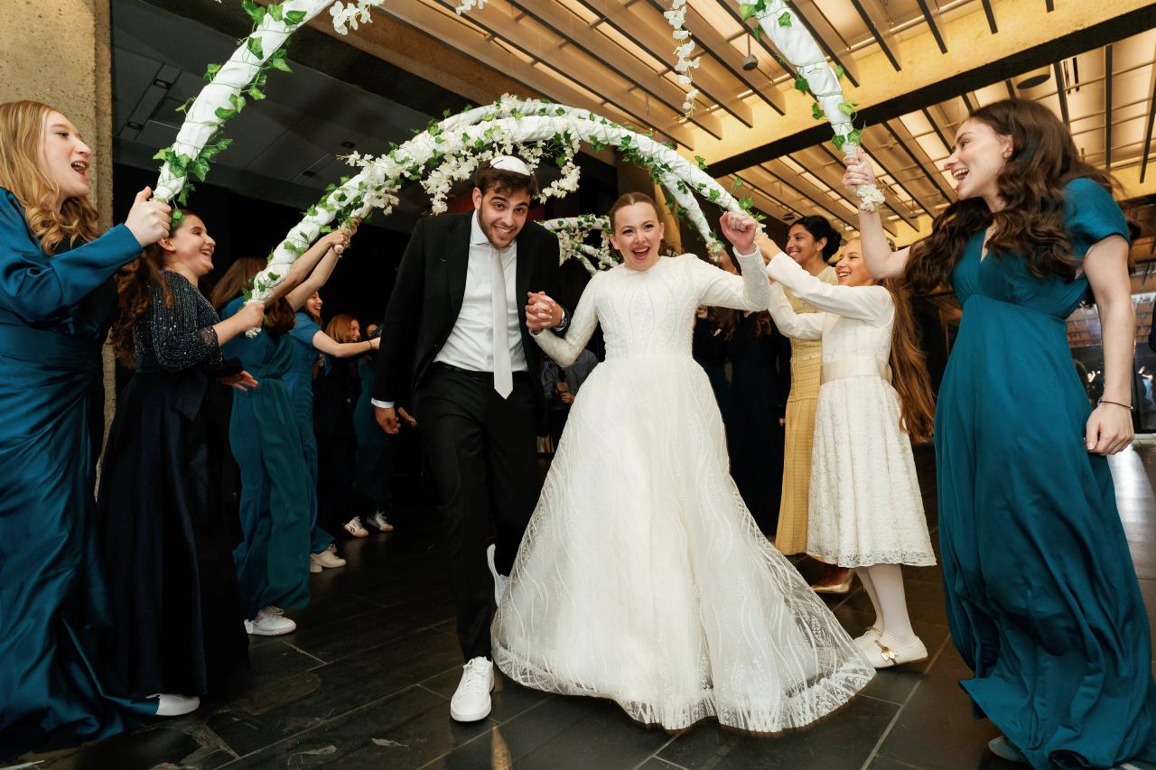 Bride and groom walking forward through a chuppah-style floral arch as guests on both sides cheer and raise their hands, with the bride’s gown billowing in motion. Wedding photography at Bellworks, Holmdel, NJ.