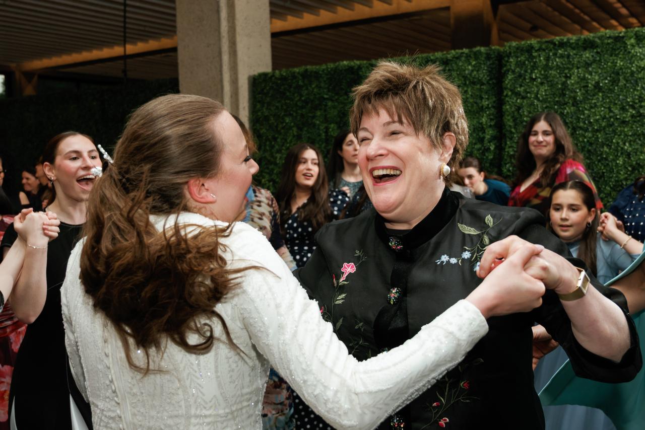 Two guests in a joyful embrace during a dance, laughing together at an event with a greenery backdrop and crowd behind them. Wedding photography at Bellworks, Holmdel, NJ.
