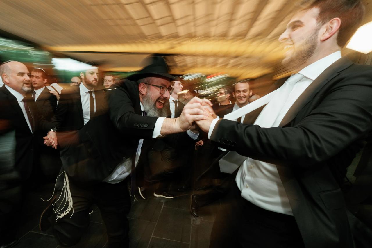 Smiling men in formalwear laughing and embracing during a lively wedding party, captured with motion blur and warm indoor lighting. Wedding photography at Bellworks, Holmdel, NJ.