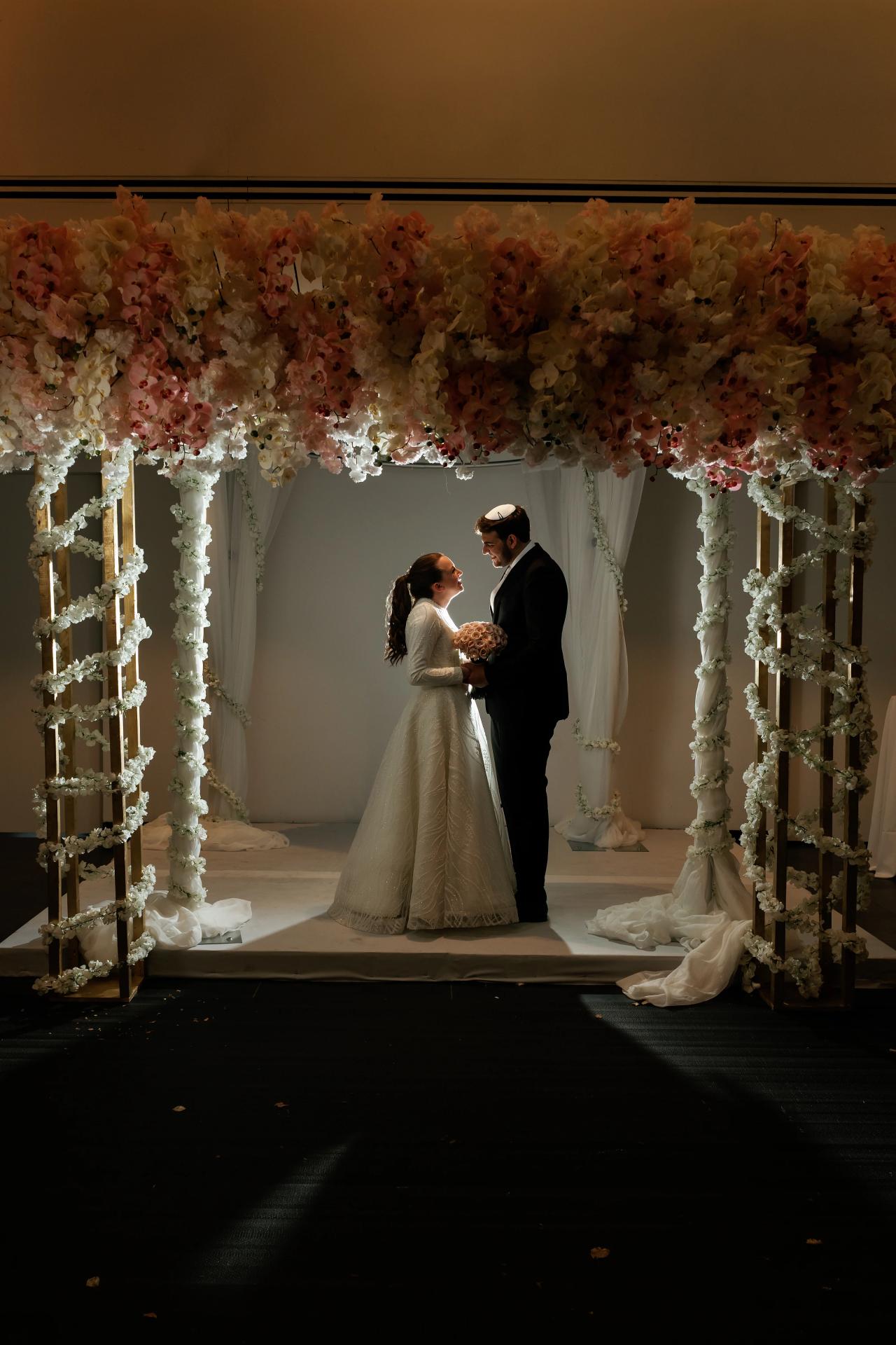 Couple standing close under an ornate floral chuppah frame during a ceremony, warm dim lighting, holding a bouquet in a quiet romantic moment. Wedding photography at Bellworks, Holmdel, NJ.