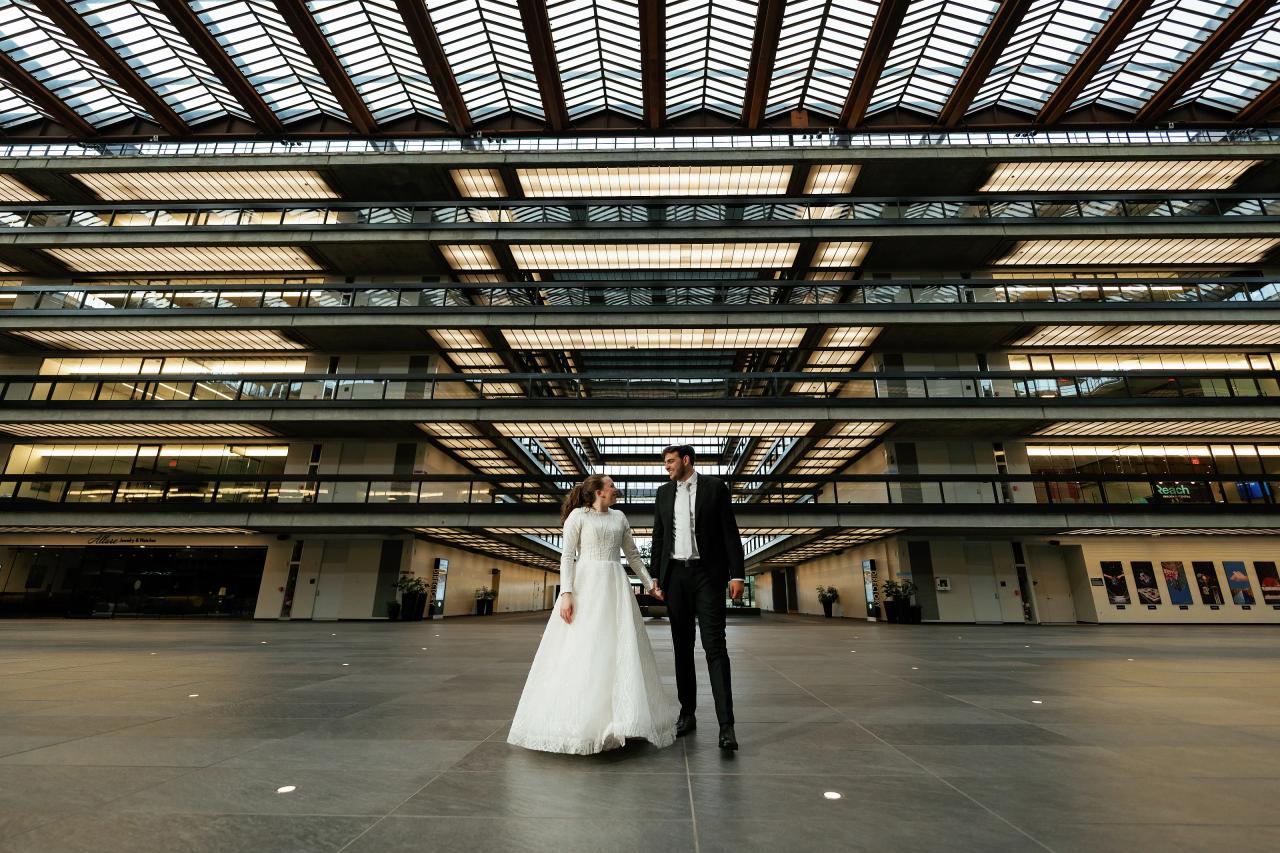 Bride in a white gown and groom in a dark suit holding hands and walking in a large modern indoor atrium with skylight ceiling panels. Wedding photography at Bellworks, Holmdel, NJ.