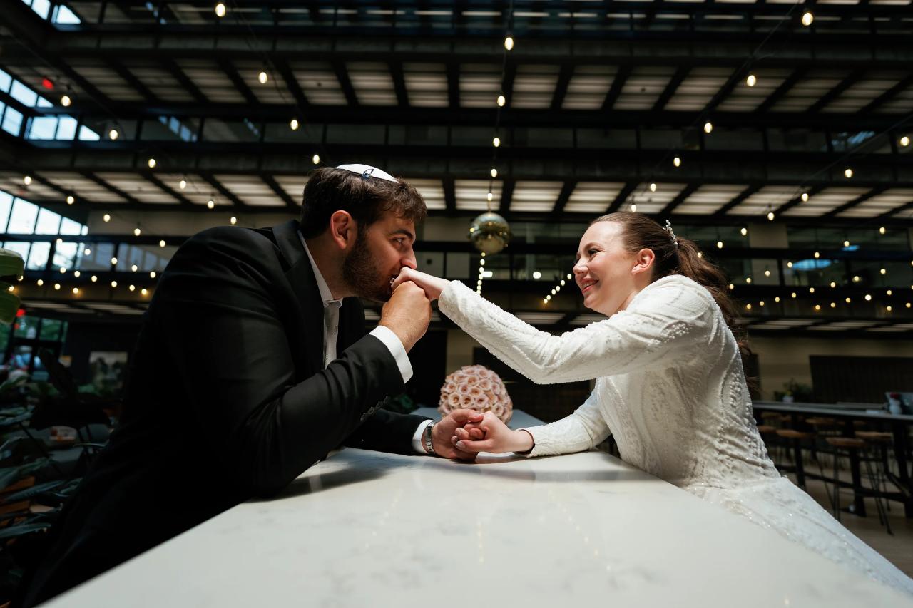 Bride in a white dress and groom in a dark suit share an intimate whisper while touching hands over a reception table, surrounded by warm string lights in an indoor hall. Wedding photography at Bellworks, Holmdel, NJ.