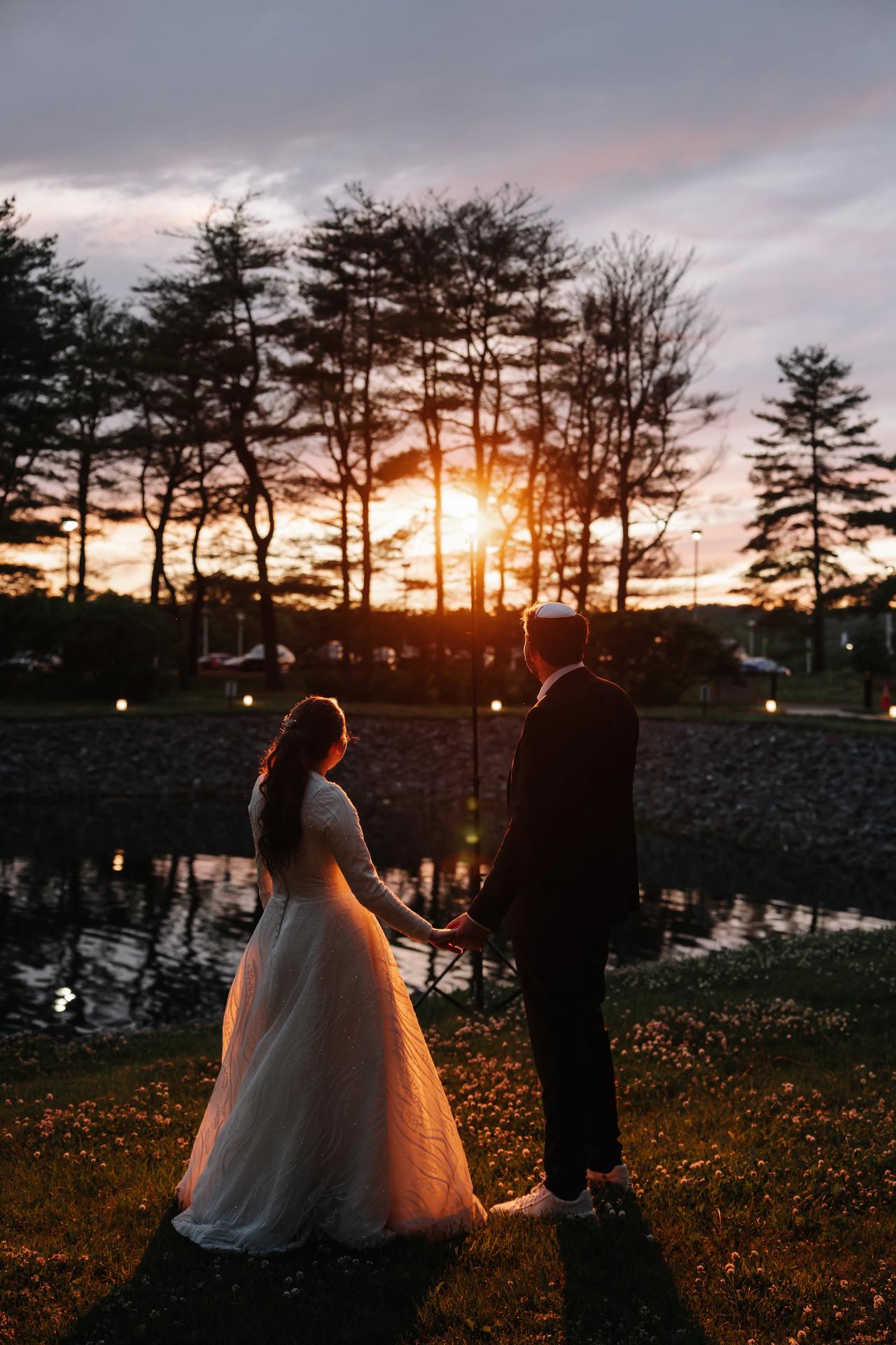 A bride and groom hold hands on a grassy path beside a pond at sunset, silhouetted against warm golden backlight with trees in the background. Wedding photography at Bellworks, Holmdel, NJ.