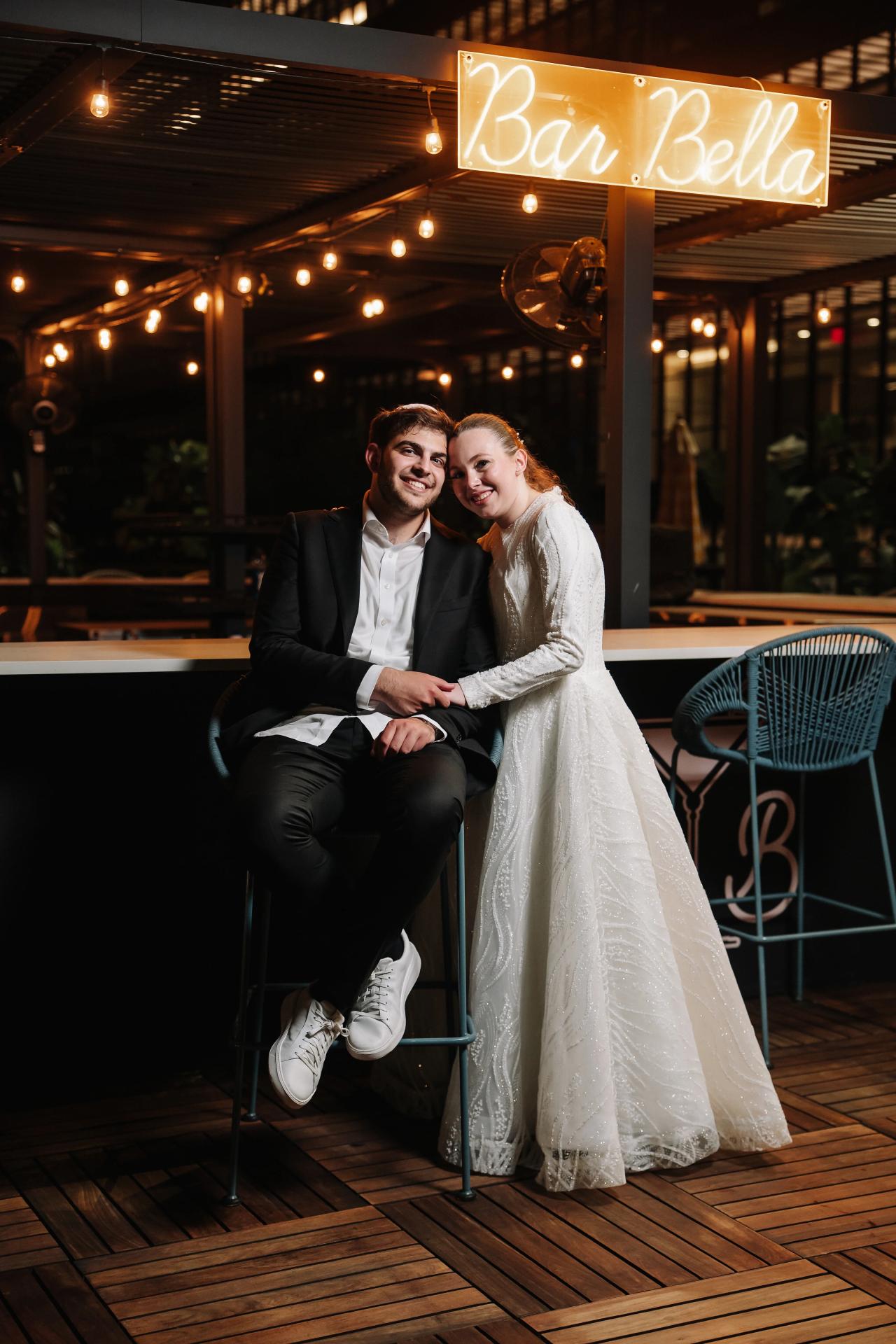 Smiling bride and groom seated close together at a warmly lit venue entrance, with the “Bar Bella” sign glowing behind them. Wedding photography at Bellworks, Holmdel, NJ.