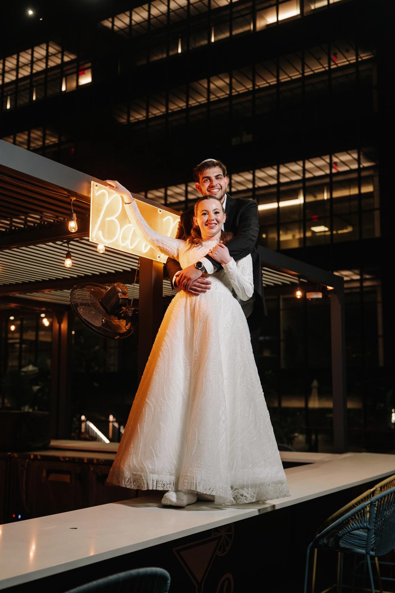 Newlyweds embracing on a reception stage under warm lights, with a glowing banner and dark industrial ceiling in the background. Wedding photography at Bellworks, Holmdel, NJ.