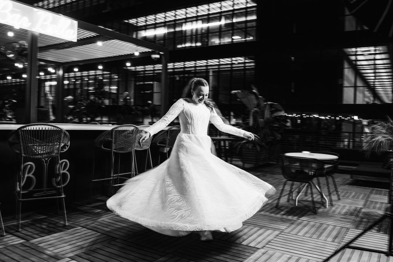 Black-and-white photo of a bride dancing mid-spin indoors at a reception, gown flowing as she turns, with chairs and lounge seating behind her. Wedding photography at Bellworks, Holmdel, NJ.