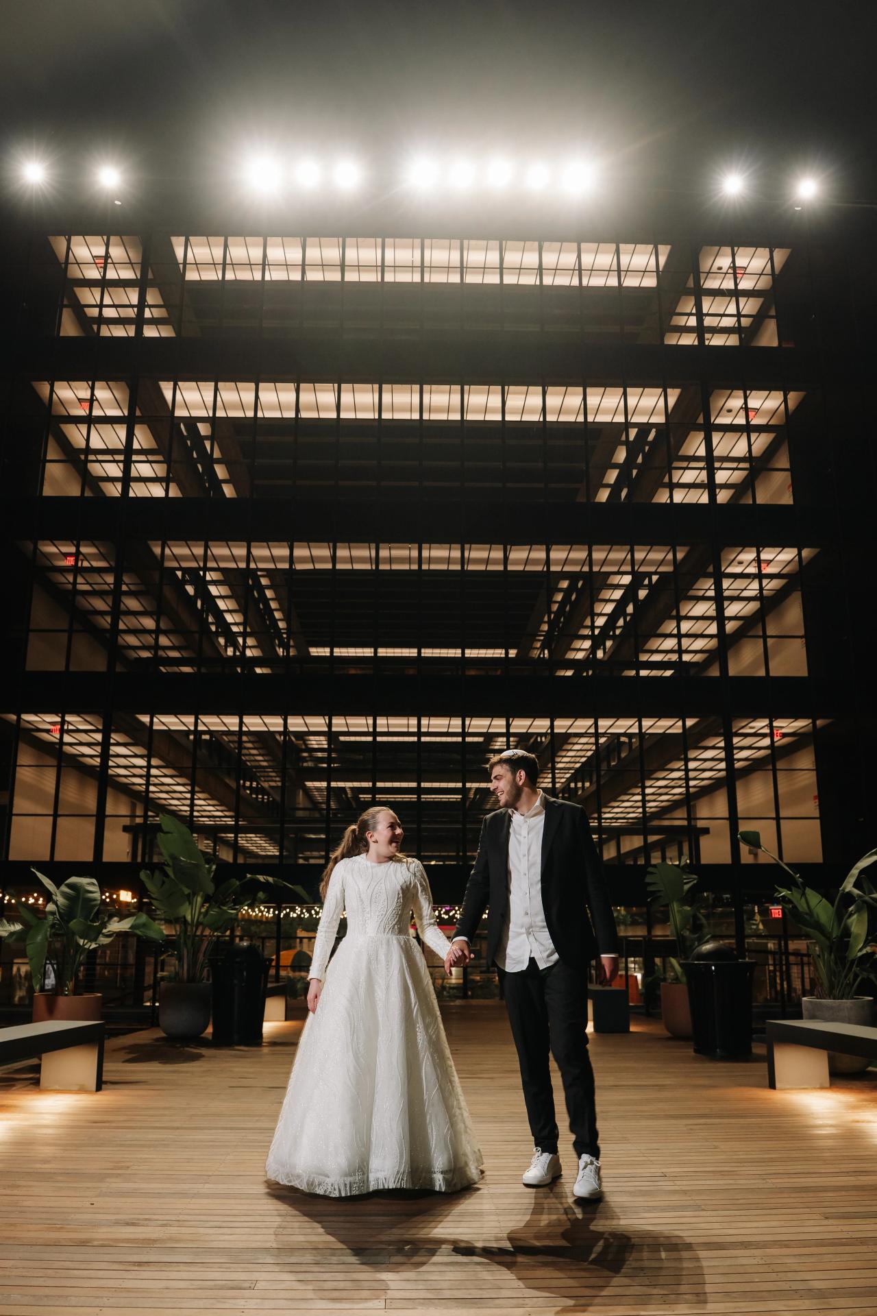 Wedding couple walking hand-in-hand inside a modern indoor venue under dramatic ceiling beams and warm overhead lights. Wedding photography at Bellworks, Holmdel, NJ.
