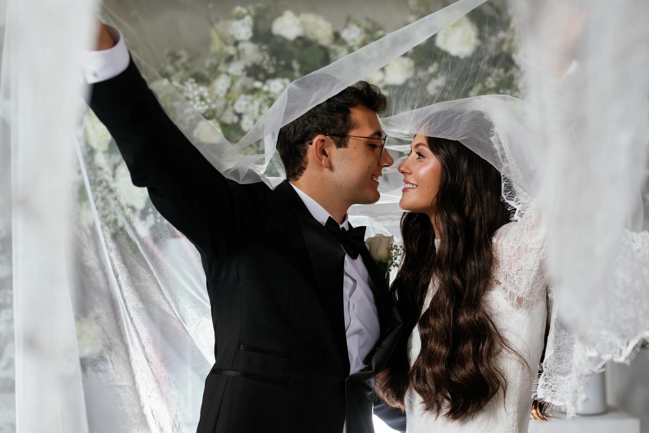 Close-up of a bride and groom under a white veil, smiling at each other during the badeken with soft light and floral backdrop behind them. Wedding photography at Falkirk Estate Country Club, Central Valley, NY.