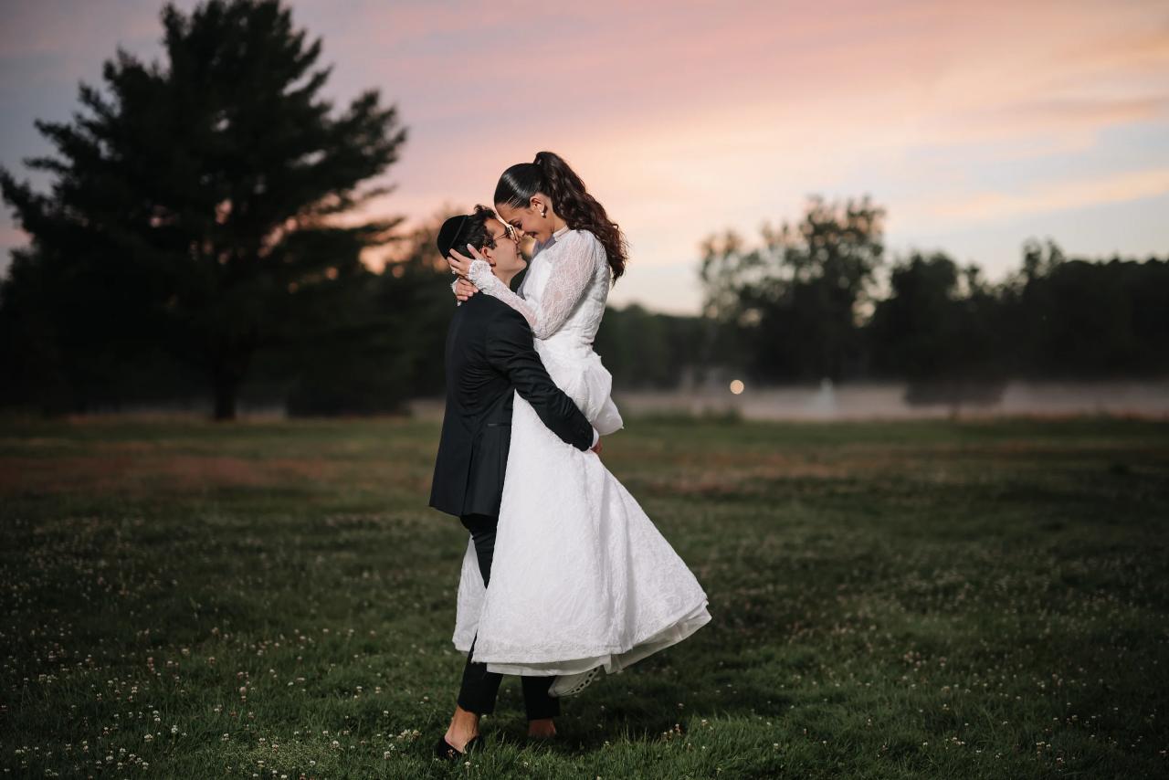 Bride and groom sharing a close kiss and embrace outdoors at golden hour in a grassy field, with a warm sunset sky and trees in the background. Wedding photography at Falkirk Estate Country Club, Central Valley, NY.