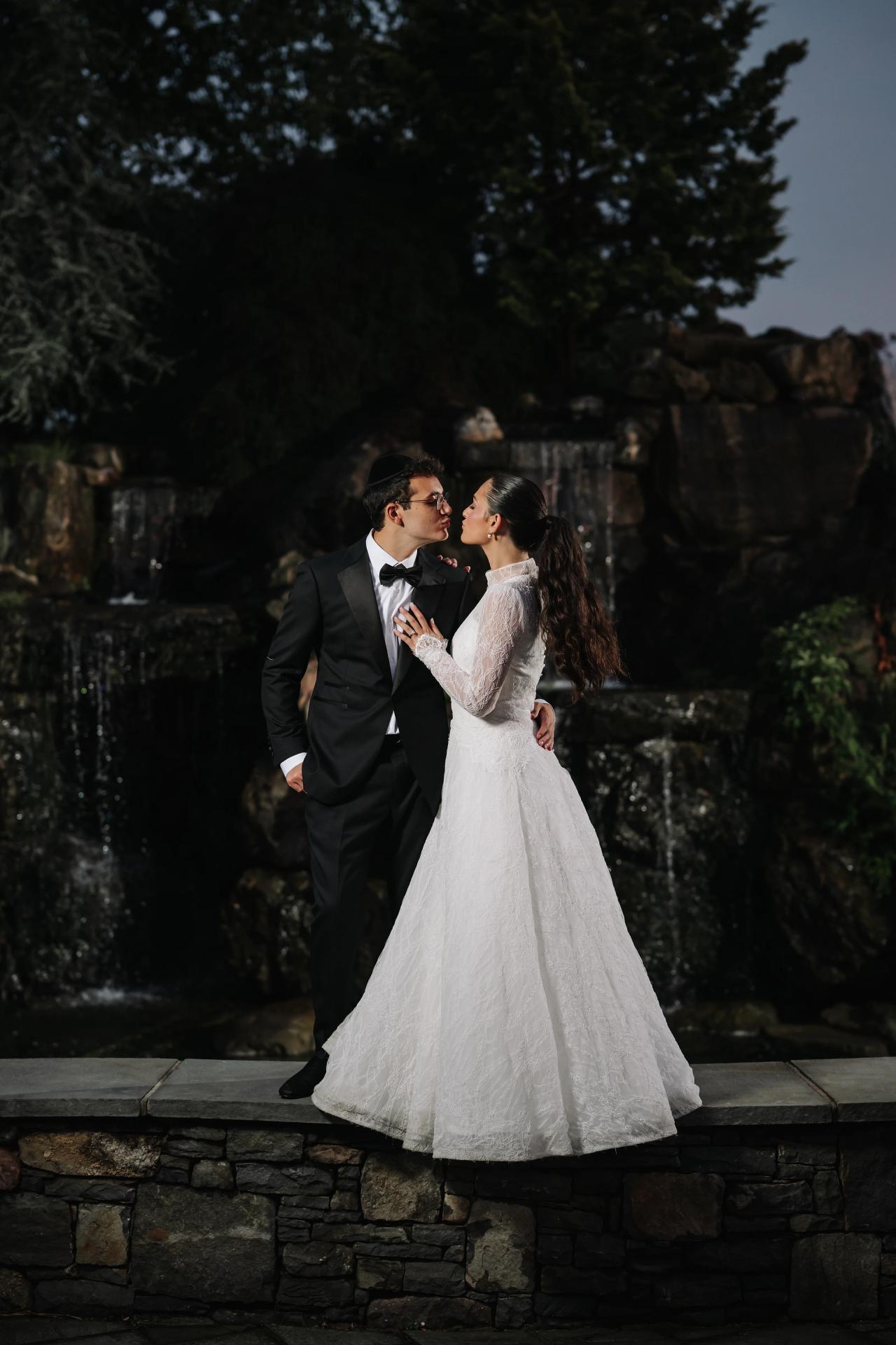 Bride and groom kissing in an intimate embrace outdoors at night, framed by a dark stone wall and greenery. Wedding photography at Falkirk Estate Country Club, Central Valley, NY.