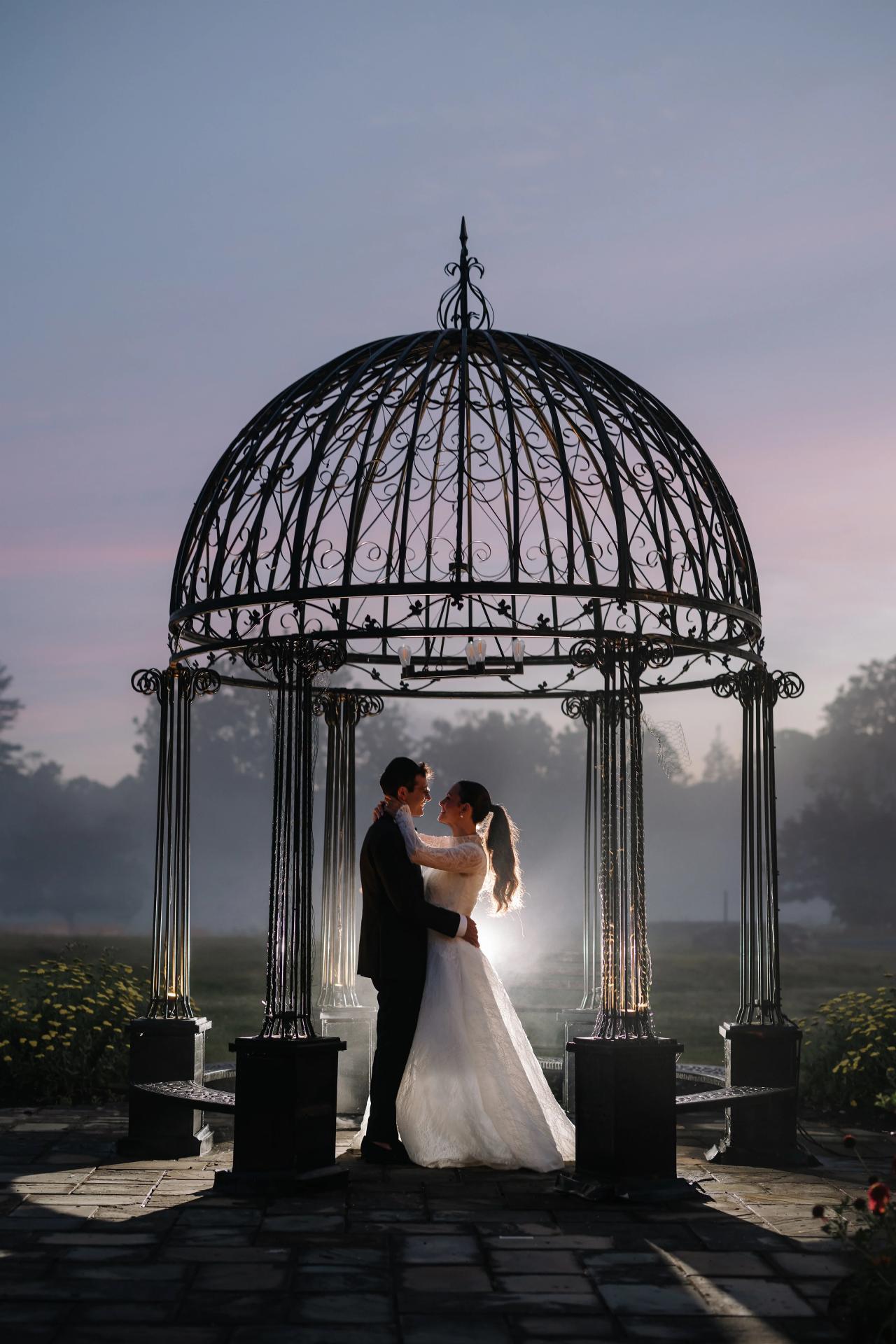 Silhouetted bride and groom standing under an ornate chuppah canopy at dusk, framed by sunset light and a misty outdoor setting. Wedding photography at Falkirk Estate Country Club, Central Valley, NY.