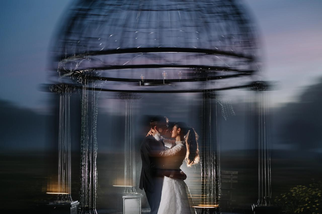A bride and groom kissing in a romantic indoor scene while water ribbons or fountain-like strands cascade around them, with an overhead canopy creating motion-blurred light. Wedding photography at Falkirk Estate Country Club, Central Valley, NY.