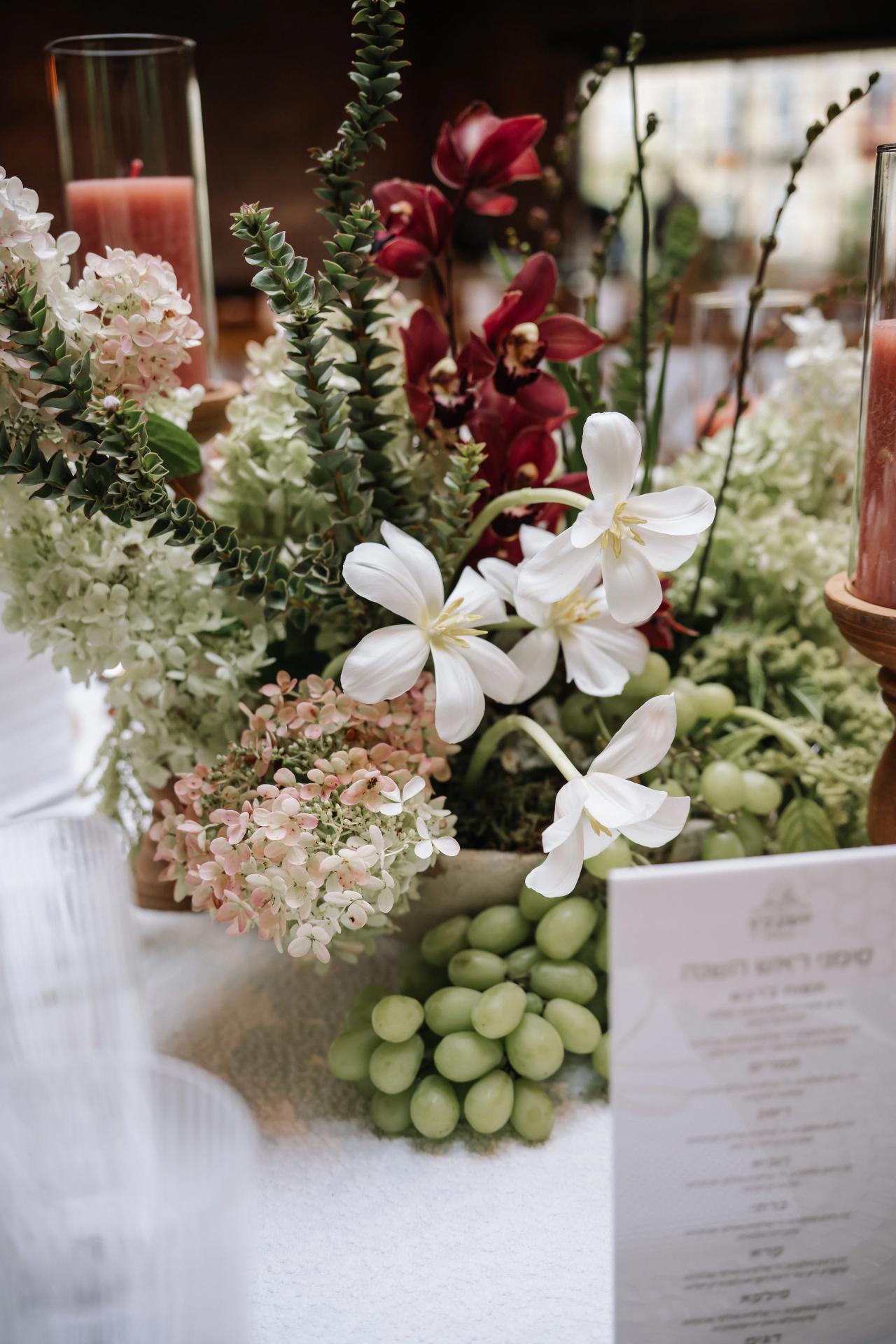 Close-up floral arrangement with white flowers, greenery, and softly blurred candles at a Bar Mitzvah reception. Bar Mitzvah photography at The Arches, Brooklyn, NY.