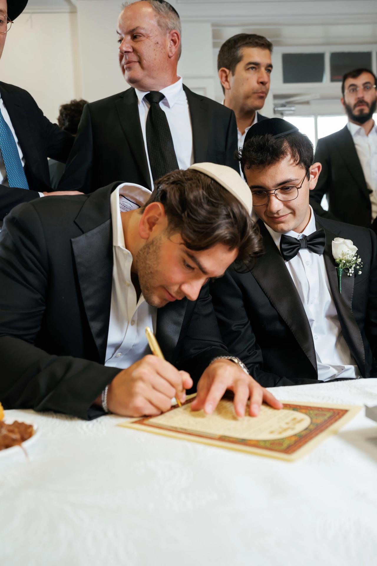 A groom in a black suit sits at a table writing carefully on a decorative document during a Jewish ritual, with family visible behind him. Wedding photography at Falkirk Estate Country Club, Central Valley, NY.