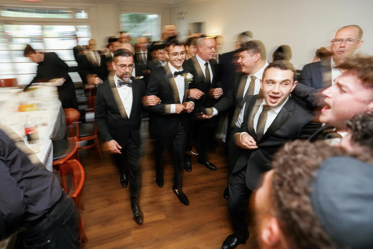 Group of men in black-tie attire laughing and moving during a wedding celebration, shot with strong motion blur in an indoor room. Wedding photography at Falkirk Estate Country Club, Central Valley, NY.