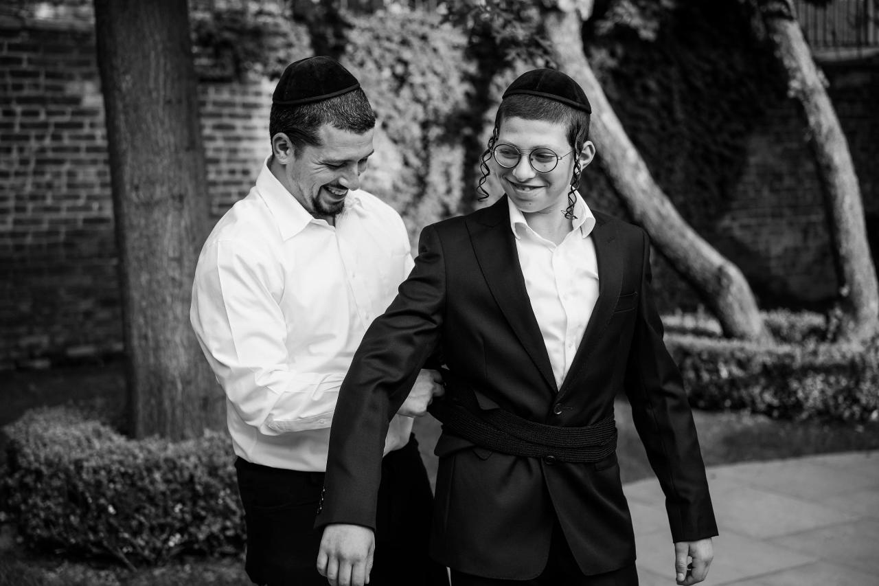 Black-and-white candid of the Bar Mitzvah boy and a guest laughing and greeting each other outdoors along a brick path with hedges in the background. Bar Mitzvah photography at The Arches, Brooklyn, NY.