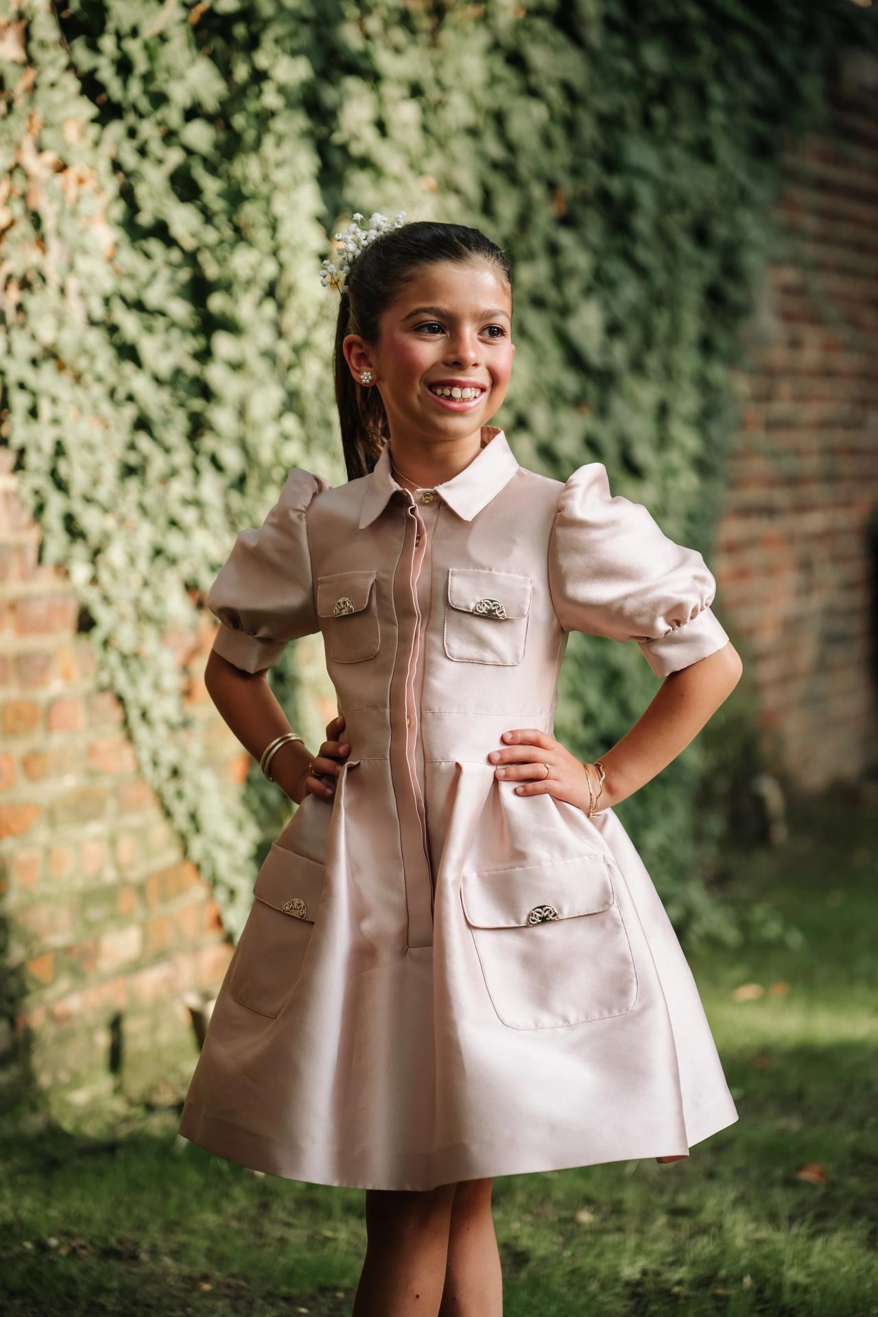 Smiling young guest outdoors in a light dress, standing with hands on hips near a brick wall and greenery. Bar Mitzvah photography at The Arches, Brooklyn, NY.