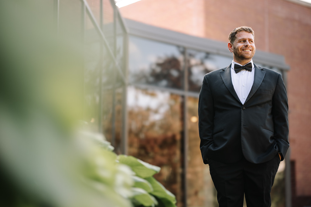 Outdoor wedding portrait of the groom in a black tuxedo and bow tie, smiling in front of a building and greenery.