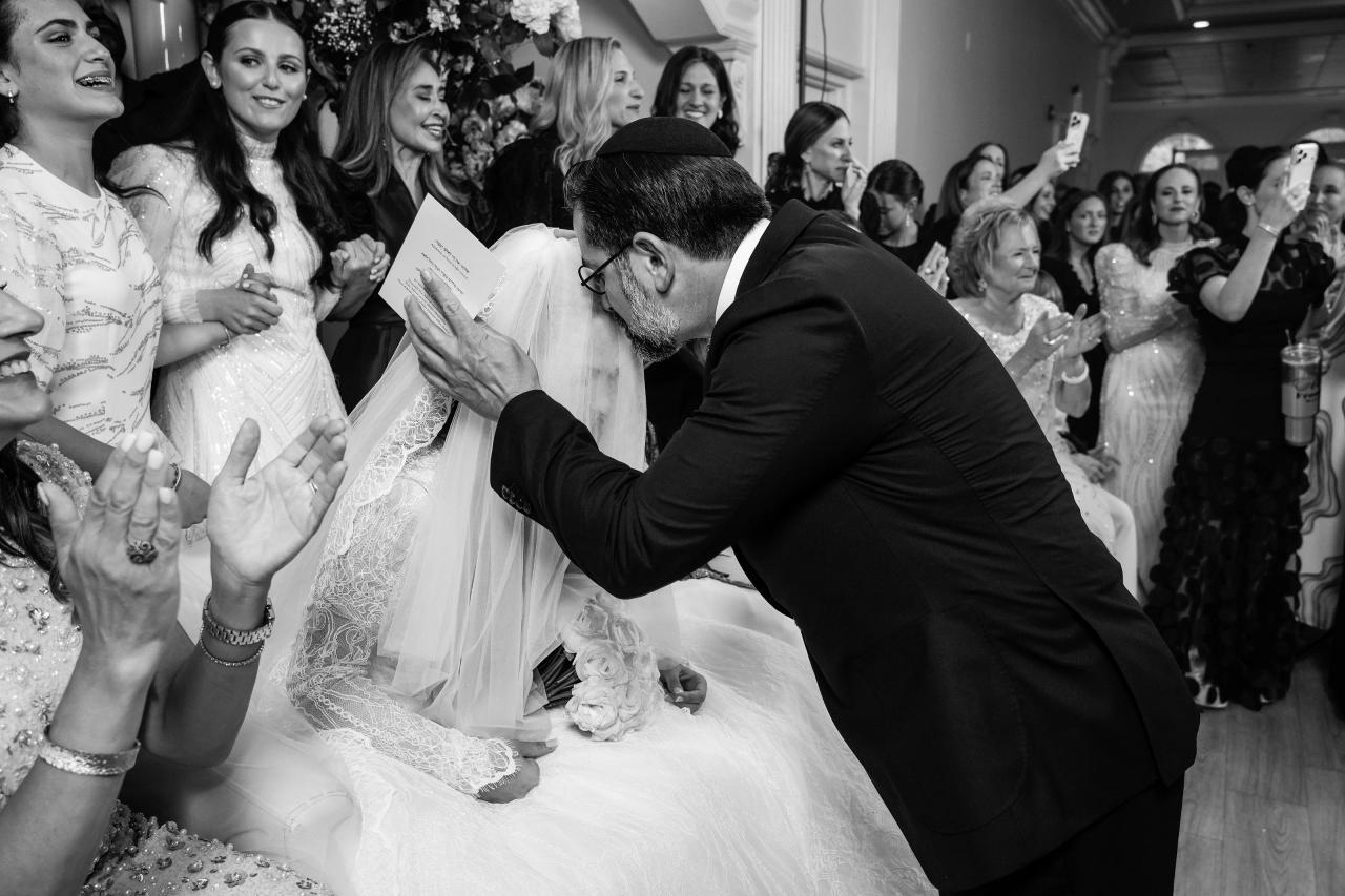 Black-and-white photo of a groom leaning in to the bride during the badeken as guests clap and watch around them. Wedding photography at Falkirk Estate Country Club, Central Valley, NY.