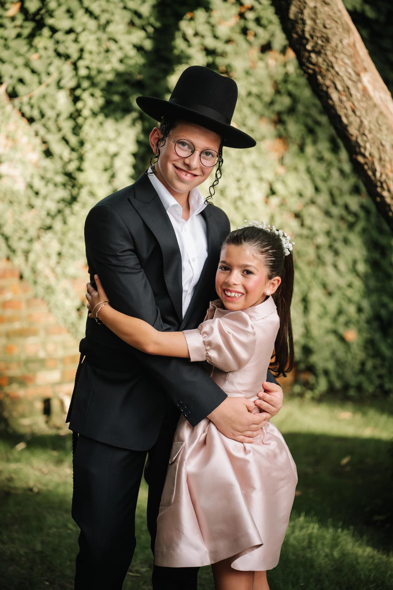 Smiling Bar Mitzvah boy and a young girl embracing outdoors, with a lush green hedge background. Bar Mitzvah photography at The Arches, Brooklyn, NY.