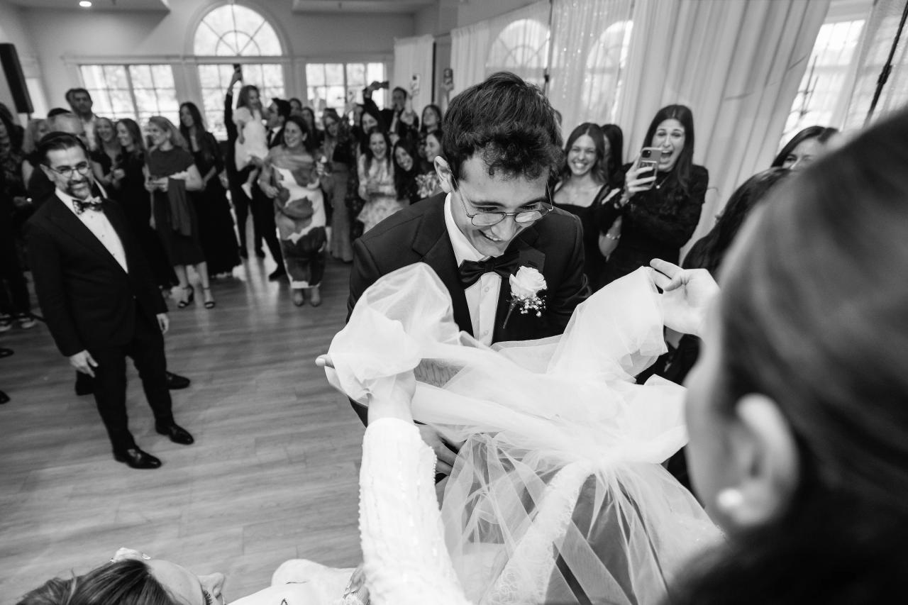 Black-and-white wedding dance-floor moment: groom in tuxedo and glasses holding a flowing white garment while guests smile and watch in the background. Wedding photography at Falkirk Estate Country Club, Central Valley, NY.