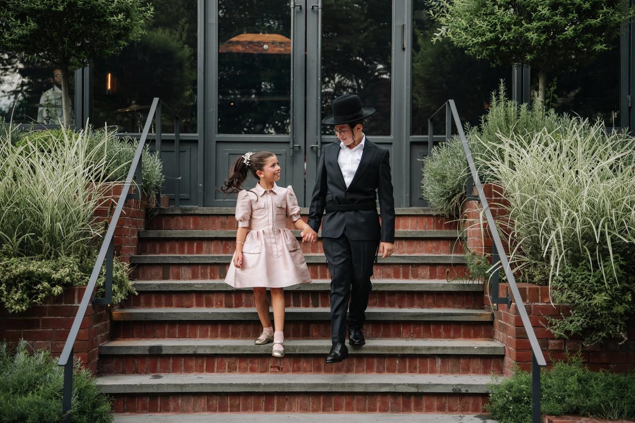 A little girl in a pale pink dress and a boy in a dark suit and black hat walk hand-in-hand up brick steps in an outdoor ceremony setting with greenery on both sides. Bar Mitzvah photography at The Arches, Brooklyn, NY.