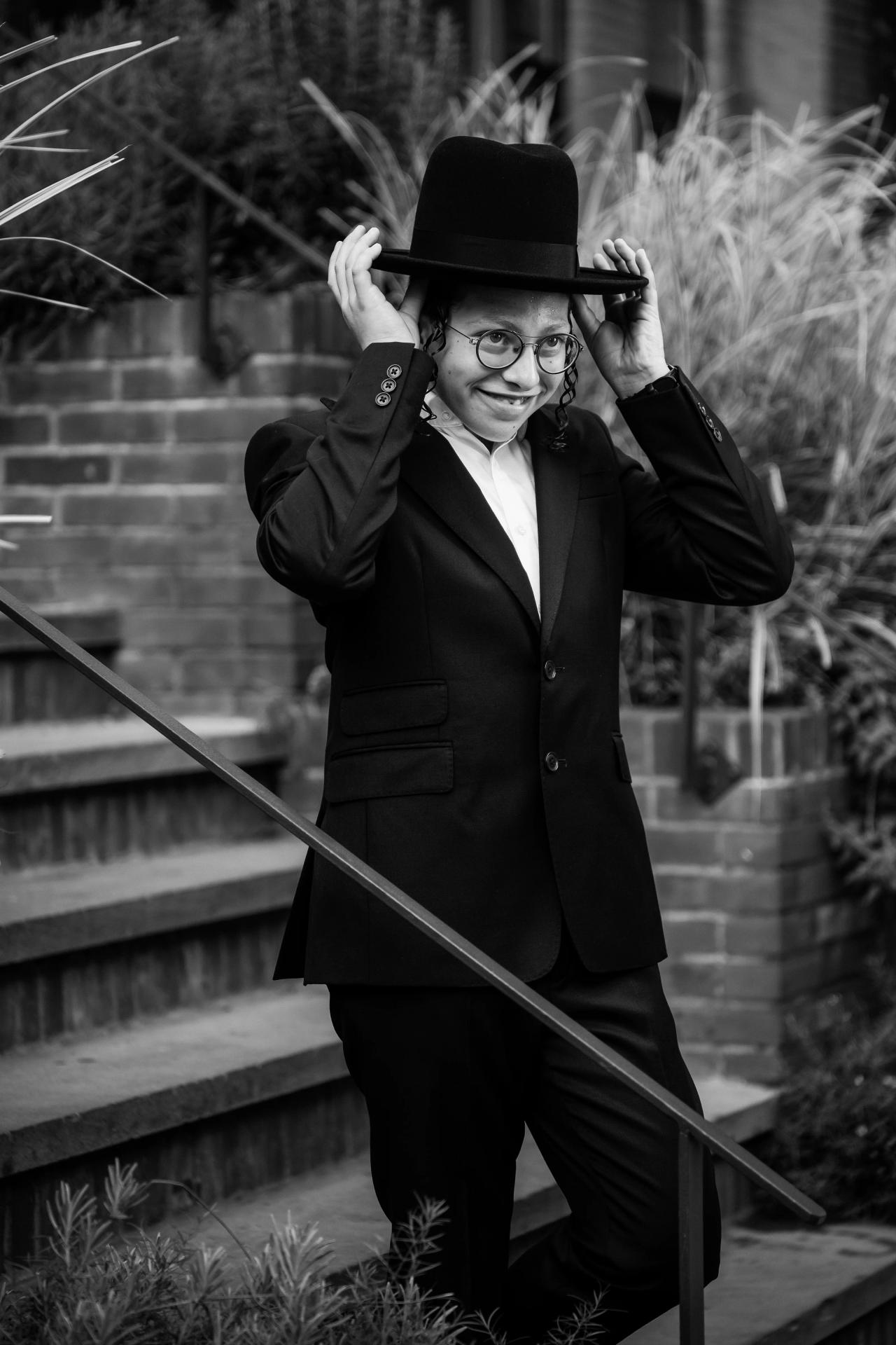 Black-and-white candid portrait of the smiling Bar Mitzvah boy holding his hat brim with both hands on a stair landing, brick wall and greenery in the background. Bar Mitzvah photography at The Arches, Brooklyn, NY.