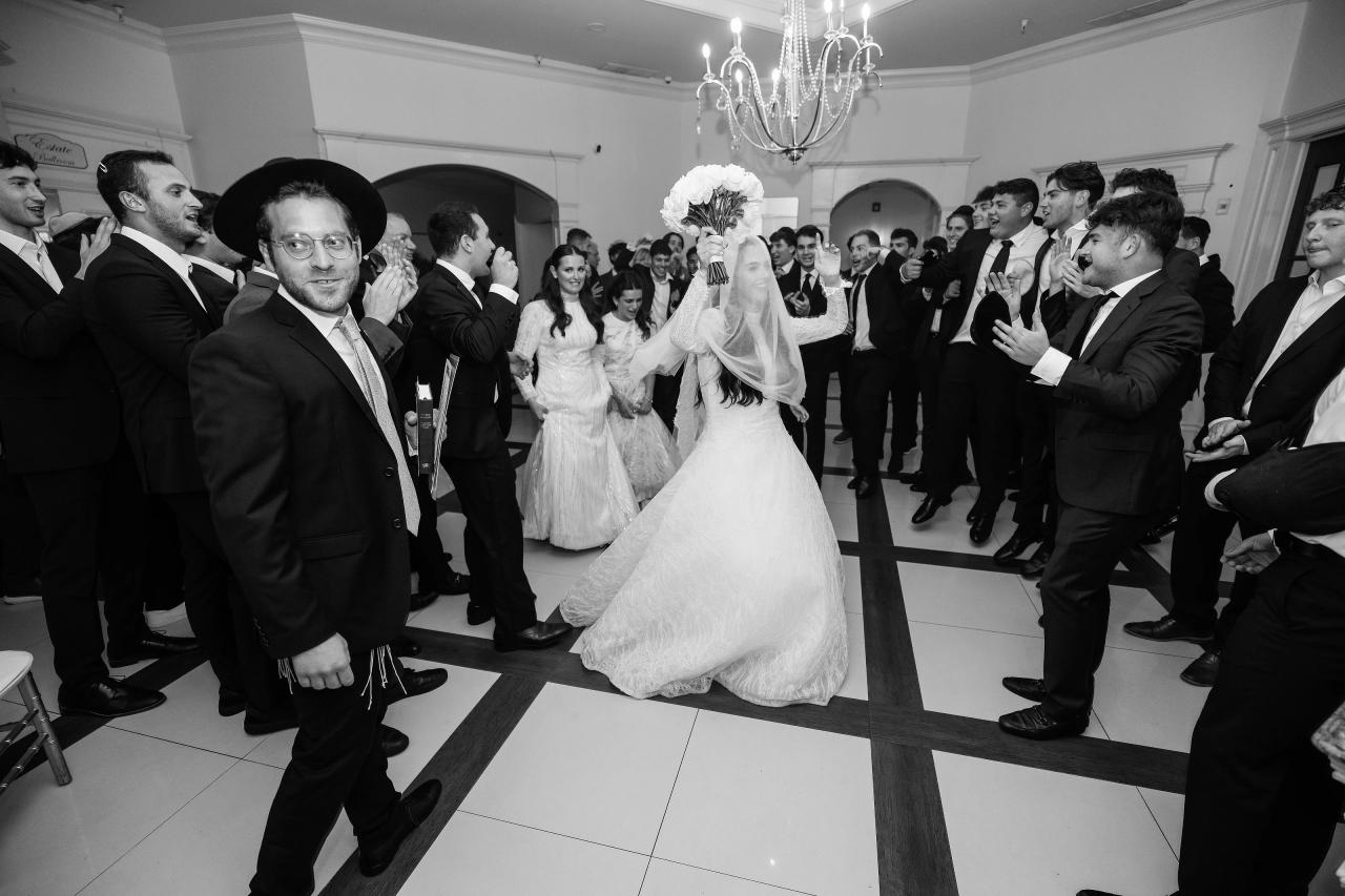 Black-and-white wedding dance-floor moment: bride in a white gown and veil walks forward through a corridor of clapping guests while the groom leads beside her. Wedding photography at Falkirk Estate Country Club, Central Valley, NY.